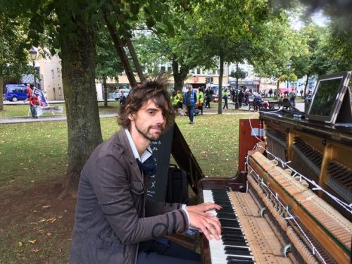 Photo of Luke Howard -- a caucasian man with brown hair and a beard in a suit, sitting at an upright piano in a public park