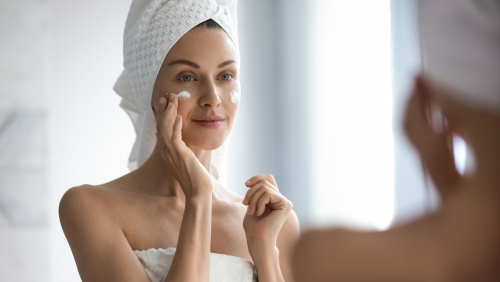 Attractive young adult woman apply facial cream look in mirror
