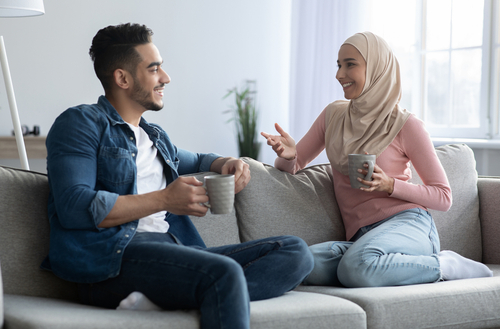 Young man and woman in hijab drinking coffee and having conversation at home. 