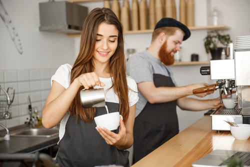  lady barista in apron preparing and pouring milk into hot cup while standing at cafe.