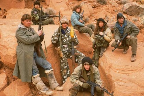 promotional image from the movie Red Dawn, featuring the cast holdinmg guns and sitting on rocks in the Utah desert