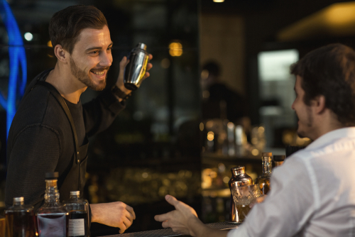 Cheerful handsome young male bartender working preparing cocktails at the bar talking to his male customer
