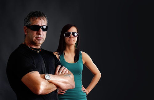 tough guy with tattoos and black sunglasses, arms crossed, looking at camera with pretty young brunette woman