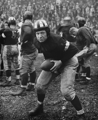 a black and white photo of a football player in a leather helmet and vintage jersey from the 1900s