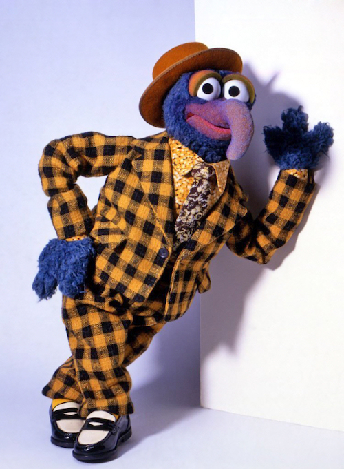 Gonzo the Great, leaning against a wall in a yellow and black checked suit with spats and a floral print shirt and fedora