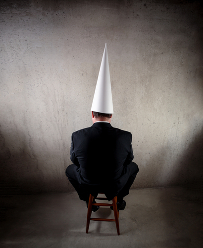 Business man wearing a dunce cap sitting on a stool with his back to the camera and facing a wall