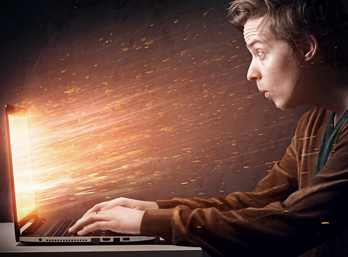 man looking with surprise at his laptop as flames and sparks leap from the screen