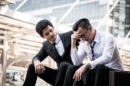 Un joven hombre de negocios asiático frustrado sentado al aire libre, con un colega poniendo una mano en su hombro para tranquilizarlo.