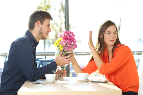 Man offering a bunch of flowers to his girlfriend in a coffee shop with an exterior background. She is holding up her hand, rejecting his offer.