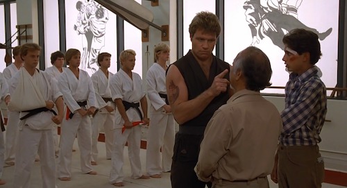 Captura de pantalla de Karate Kid. El Sr. Miyagi y Daniel Larusso confrontan a Sensei Kreese en su dojo, frente a su clase. Kreese está señalando agresivamente al Sr. Miyagi