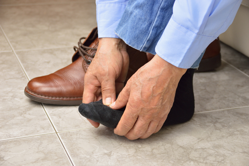 imagen recortada de los brazos de un hombre con una camisa oxford azul frotándose el pie dolorido, un zapato a su lado en el suelo