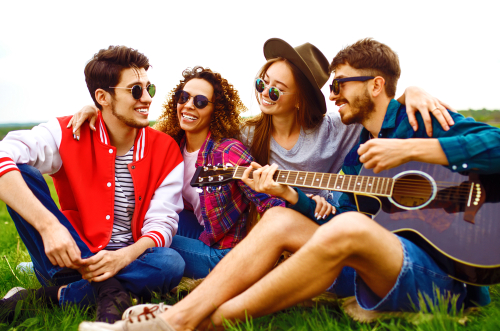 Group of happy friends with guitar having fun spending free time together in park sitting on grass