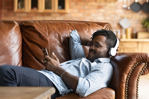 Calm young african ethnicity man in glasses wearing headphones, relaxing on comfortable couch listening favorite music
