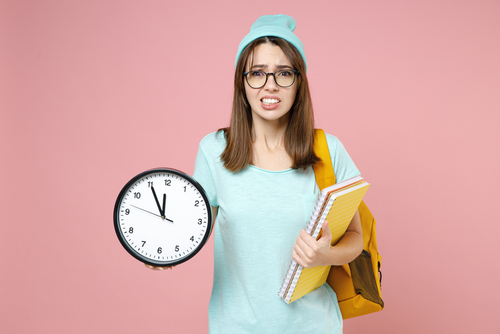 Preocupada joven estudiante en casual camiseta azul sombrero gafas mochila sujetar en manos cuadernos reloj aislado sobre fondo de color rosa pastel