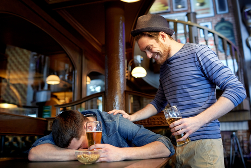 man with beer waking his drunk friend sleeping on table at bar 