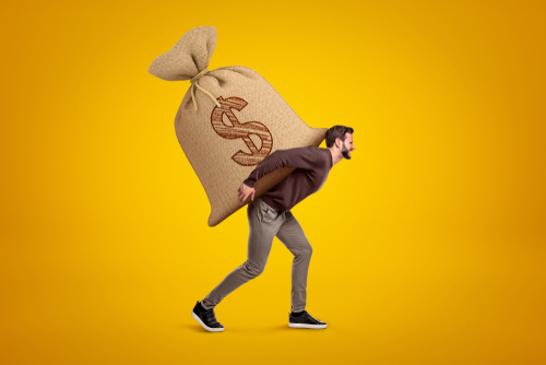 Side view of young handsome man in casual clothes carrying huge heavy sack with dollar symbol on. 