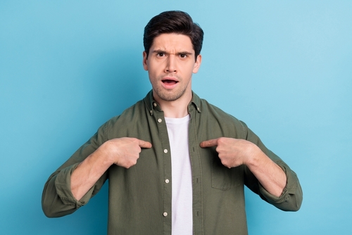 foto de un hombre blanco con una camiseta verde oliva y una camiseta blanca aislada de fondo azul, apuntándose a sí mismo con ambas manos y luciendo sorprendido