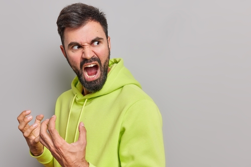Bearded, white man in neon green hoodie on grey background looks over his shoulder, gesturing angrily and yelling