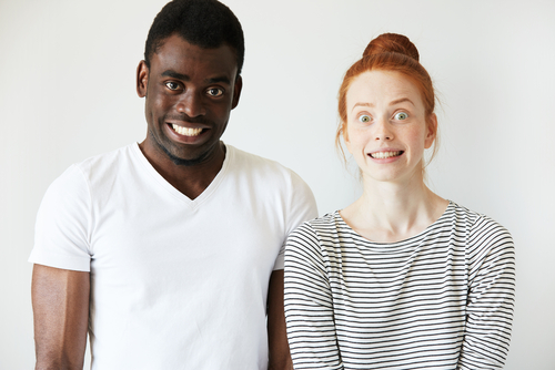 A black man and white woman isolated on a white background, standing close to one another with wide eyes and obviously fake smiles, looking tense and stressed.